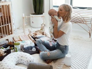 Woman sitting on the floor in a nursery holding and kissing a baby, surrounded by baby care items and toys.