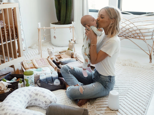 Woman sitting on the floor in a nursery holding and kissing a baby, surrounded by baby care items and toys.
