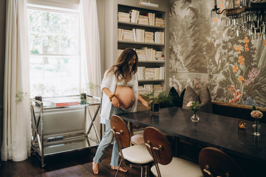 Pregnant woman standing barefoot in a dining room with bookshelves and floral wallpaper, touching her belly.