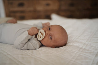Baby lying on a bed with a pacifier in mouth, looking towards the camera