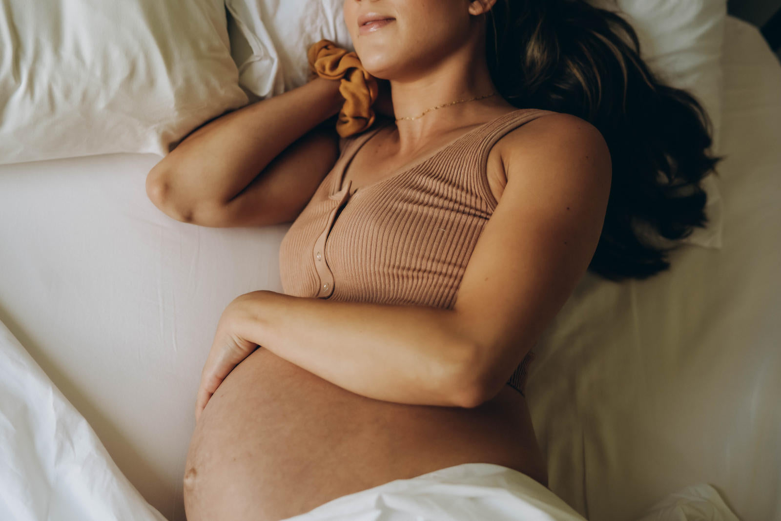 Pregnant woman lying on a bed, resting with one hand on her belly while wearing a ribbed tan tank