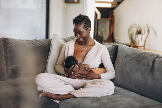 Mother breastfeeding her newborn baby while sitting cross-legged on a gray couch in a cozy living room.