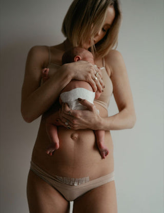 Woman holding a newborn baby against her bare torso, showing a fresh C-section scar on her abdomen.