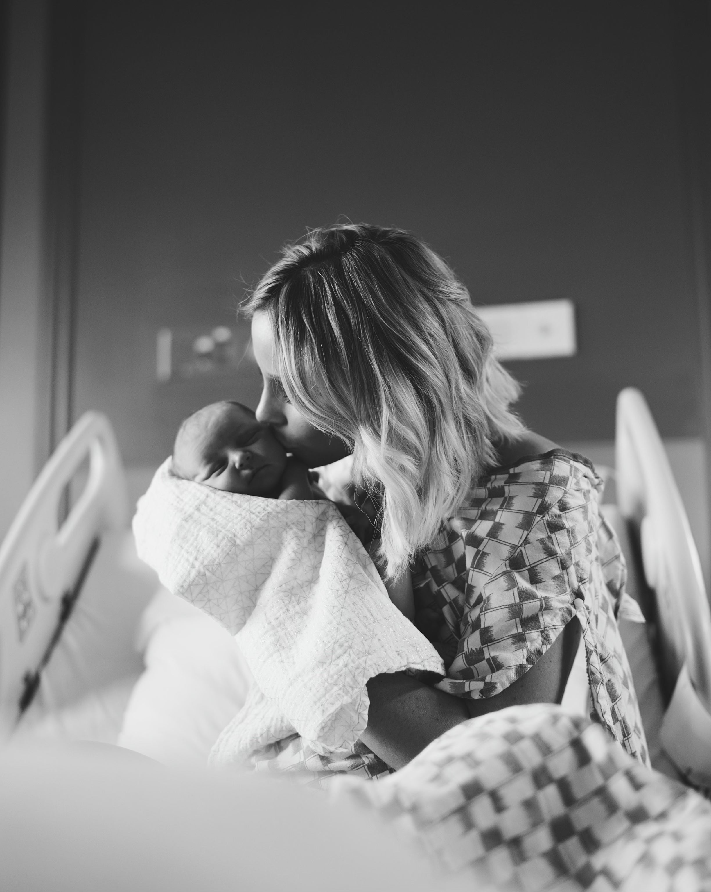 Mother holding and kissing newborn wrapped in a blanket in a hospital room