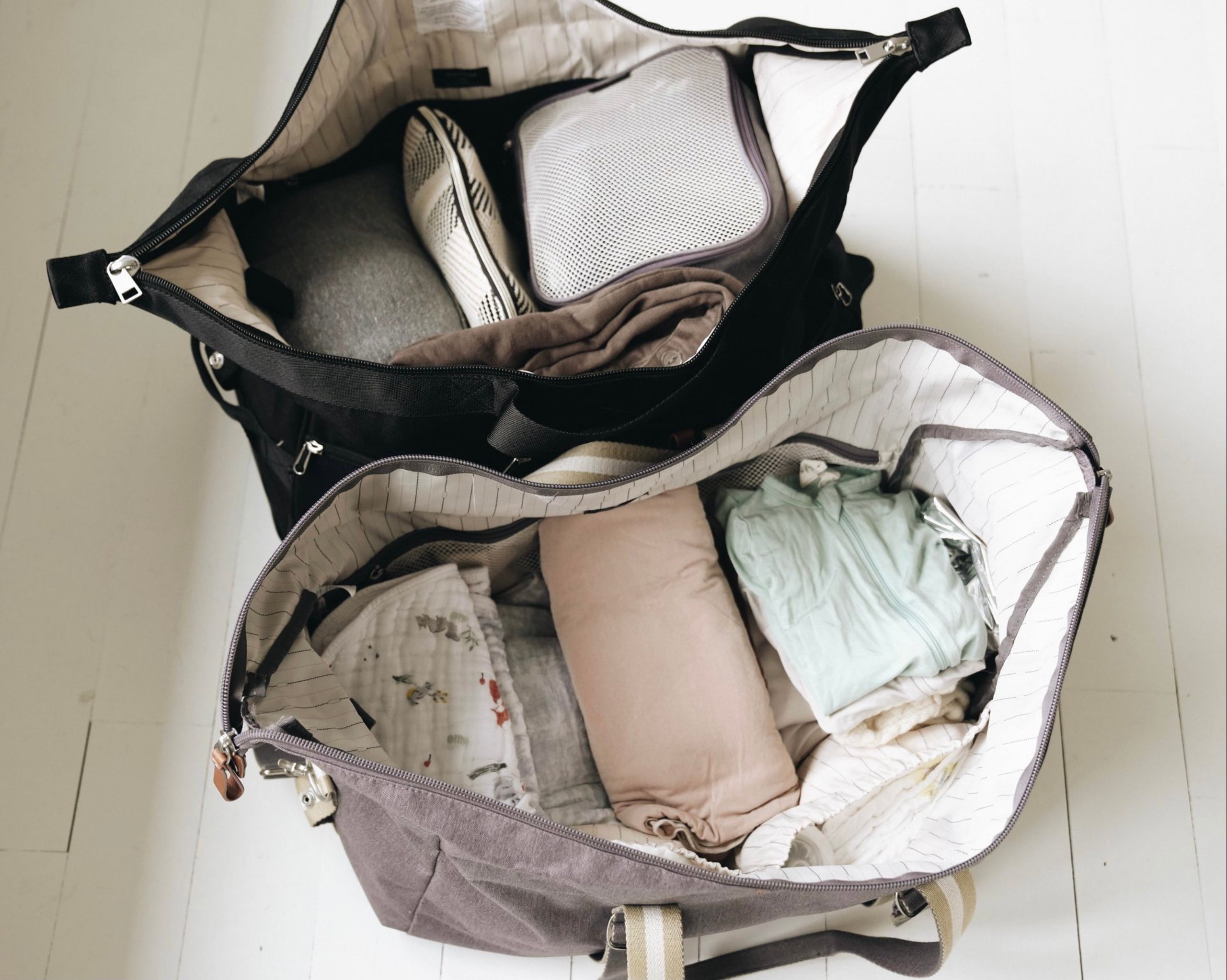 Open duffel bag packed with folded baby clothes, swaddles, a rolled pillow and a mesh packing cube on a white wooden floor