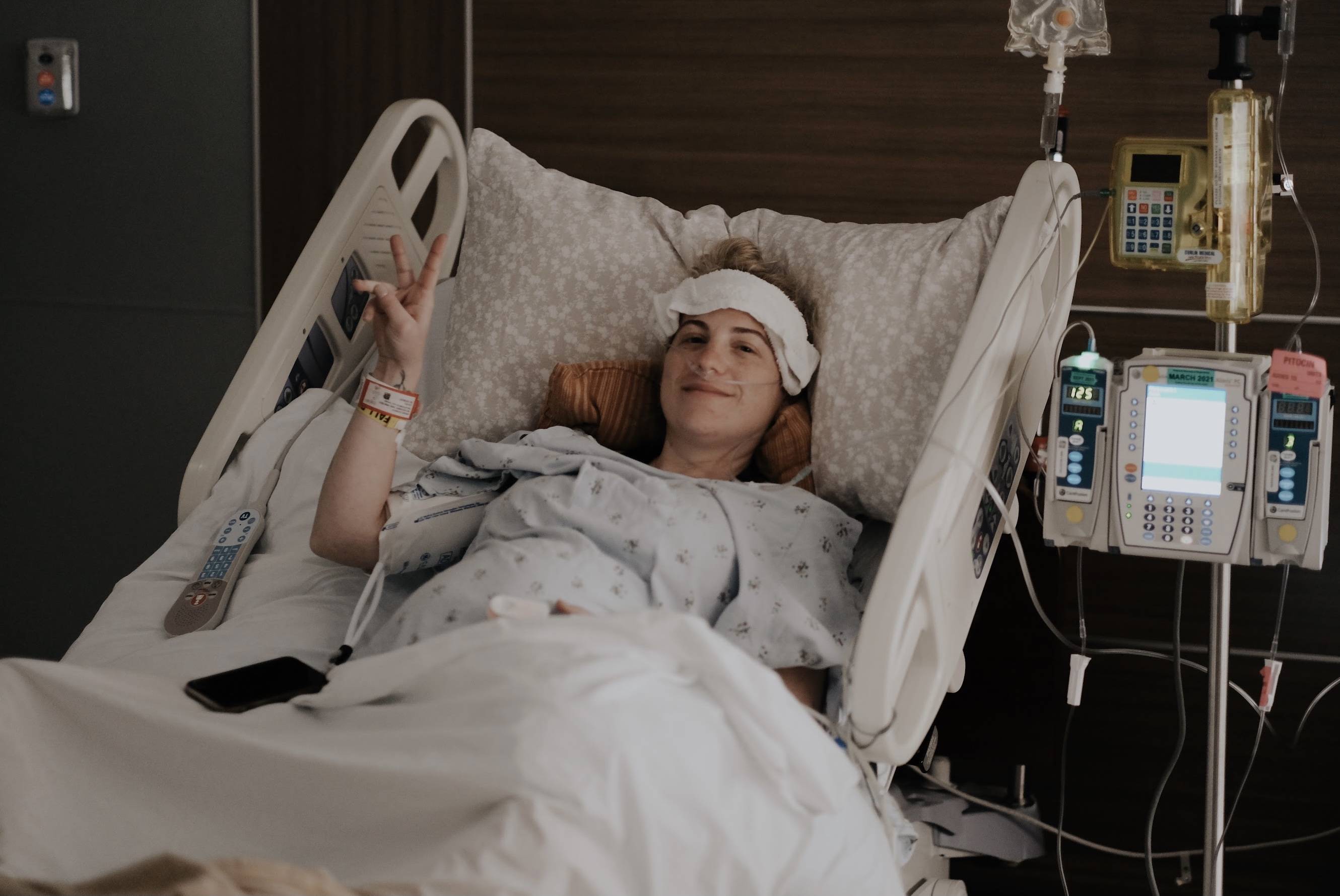 Person lying in a hospital bed giving a peace sign, wearing a forehead wrap and nasal cannula with IV pumps visible at bedside
