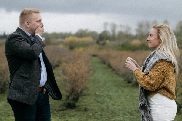 Man covering his mouth in surprise while a woman smiles at him in a shrub-lined field under an overcast sky