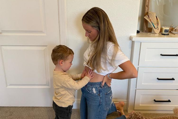 Woman kneeling while a young boy touches her abdomen beside a white dresser with jewelry on top in a bedroom-like room