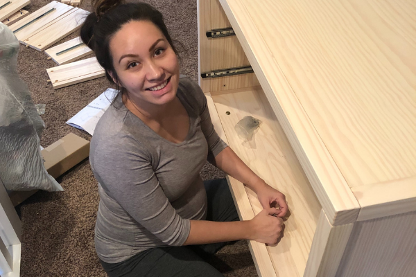 Sadie Powell kneeling and smiling while assembling a light-wood dresser in a carpeted room