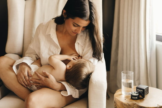Woman breastfeeding a child while sitting on a chair next to a small table with a glass of water and skincare products.