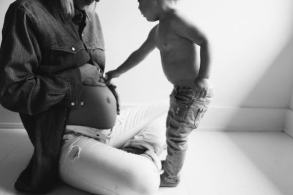Pregnant woman sitting on the floor with a young child standing and touching her belly in a softly lit room.