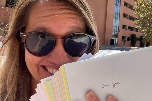 Woman wearing sunglasses smiling and holding a stack of papers, the top paper has handwritten text 'For you.'