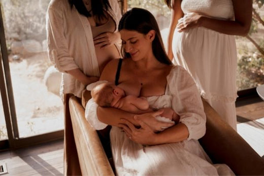 Mother breastfeeding an infant while two pregnant women stand nearby in a sunlit living room