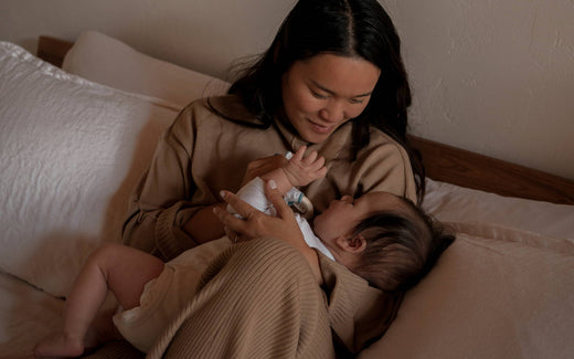 Mother bottle-feeding an infant while sitting on a bed in a warm, neutral-toned room