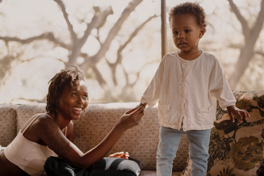 Woman smiling at a toddler standing on a couch in a sunlit room, illustrating child safety and interaction.