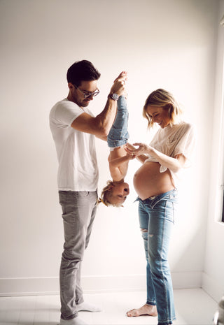 A pregnant woman smiling at a toddler held upside down by a man, all in casual clothes in a bright room.