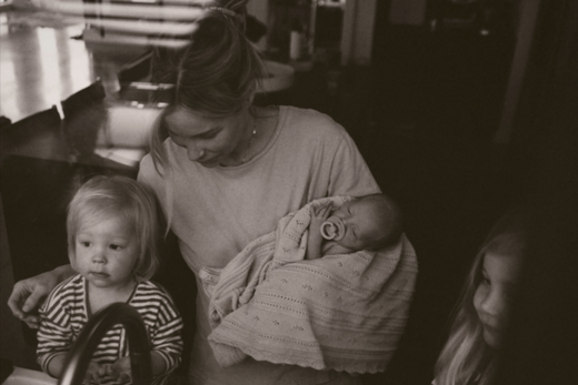 A woman holding a swaddled baby with a pacifier, sitting with two young children, one toddler and one older child, in a dimly lit room.