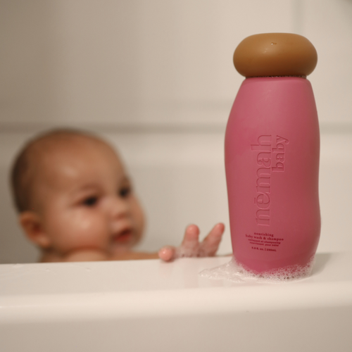 Baby reaching toward a pink Nēmah baby wash bottle beside a bathtub