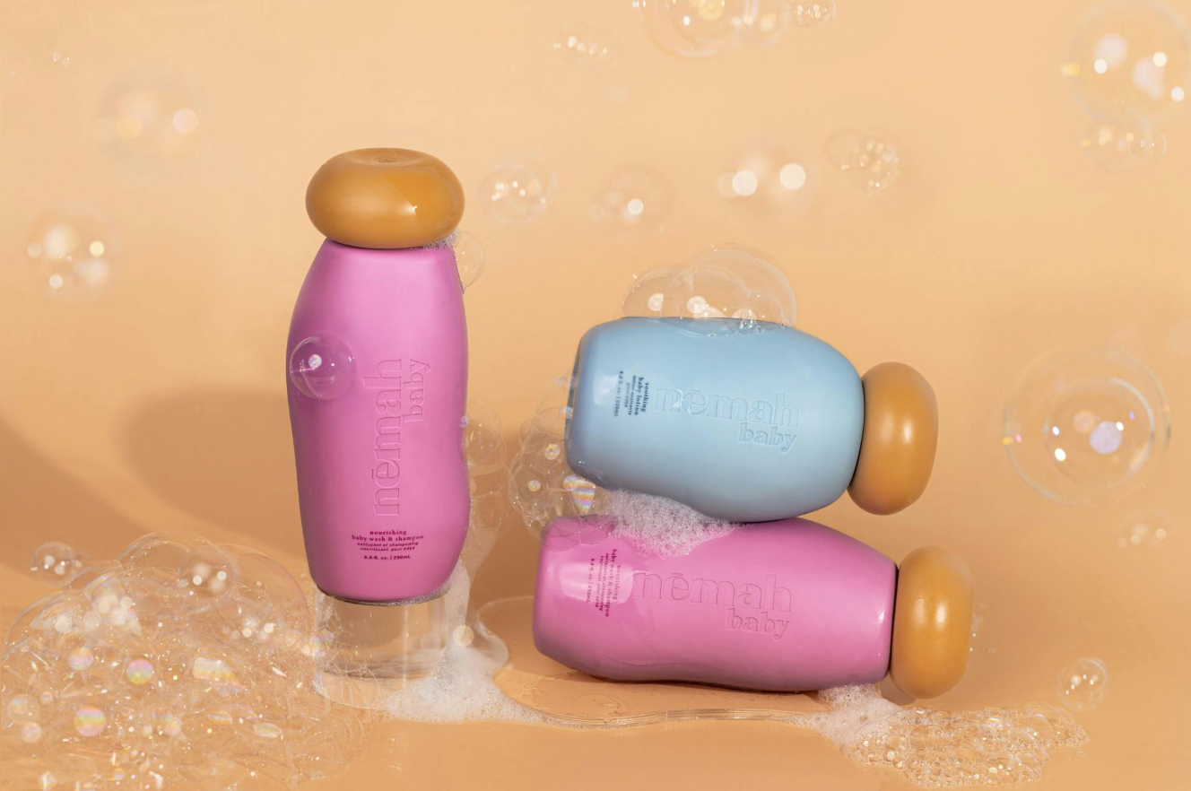 Angled view of 3 Nēmah Baby bottles showing tan twist-dispense caps and embossed logo on peach background with soap bubbles