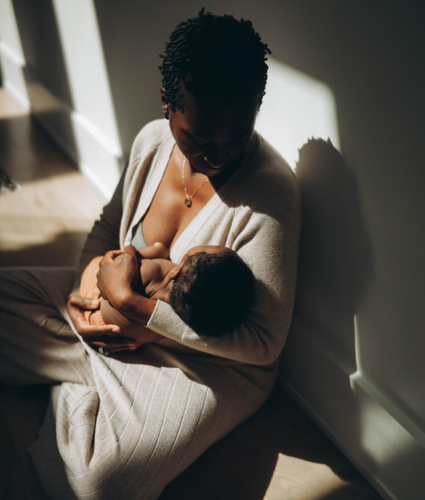 Mother breastfeeding an infant on the floor beside a sunlit wall, soft natural light creating warm shadows