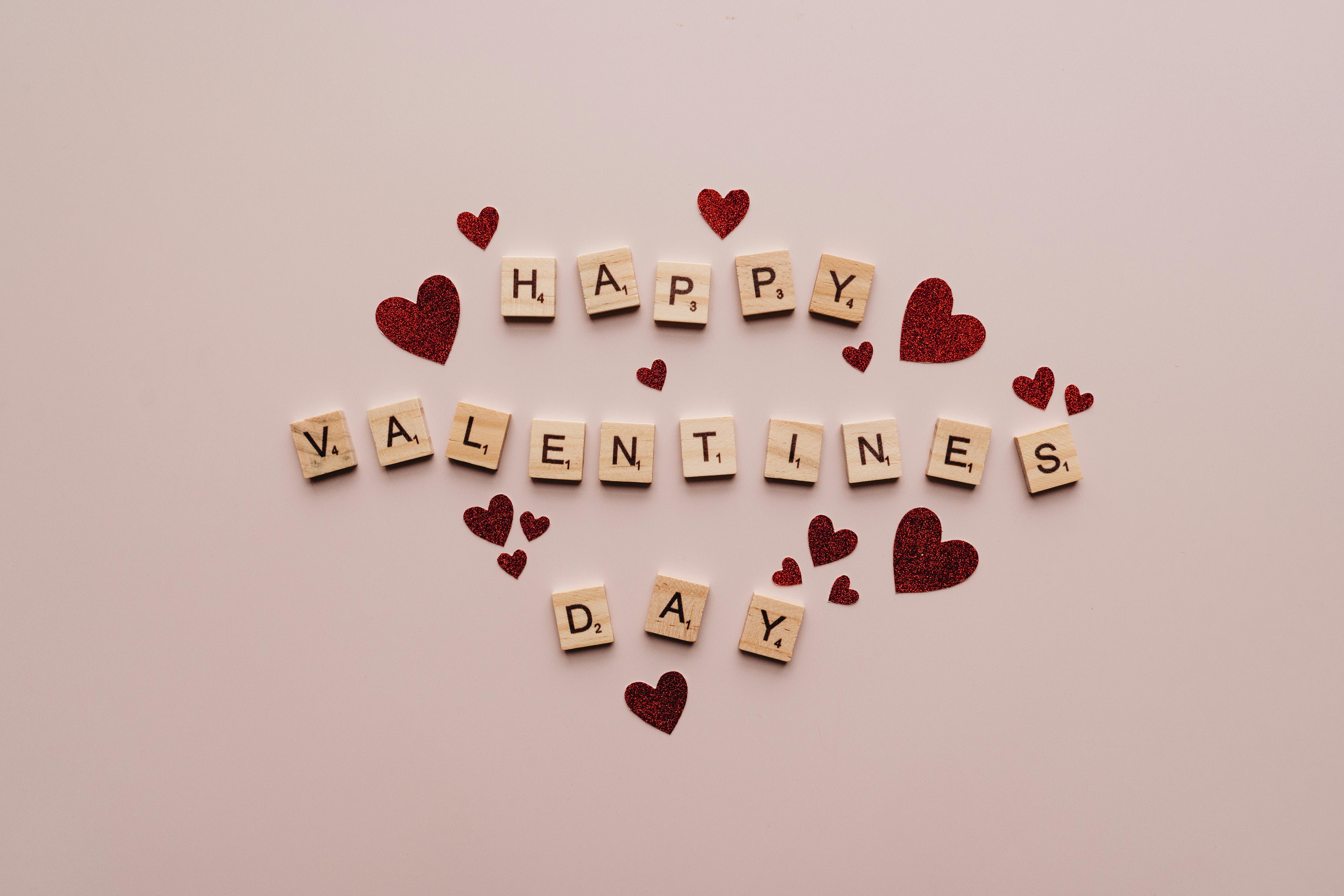 Scrabble tiles spelling Happy Valentines Day surrounded by red glitter hearts on a pale pink background