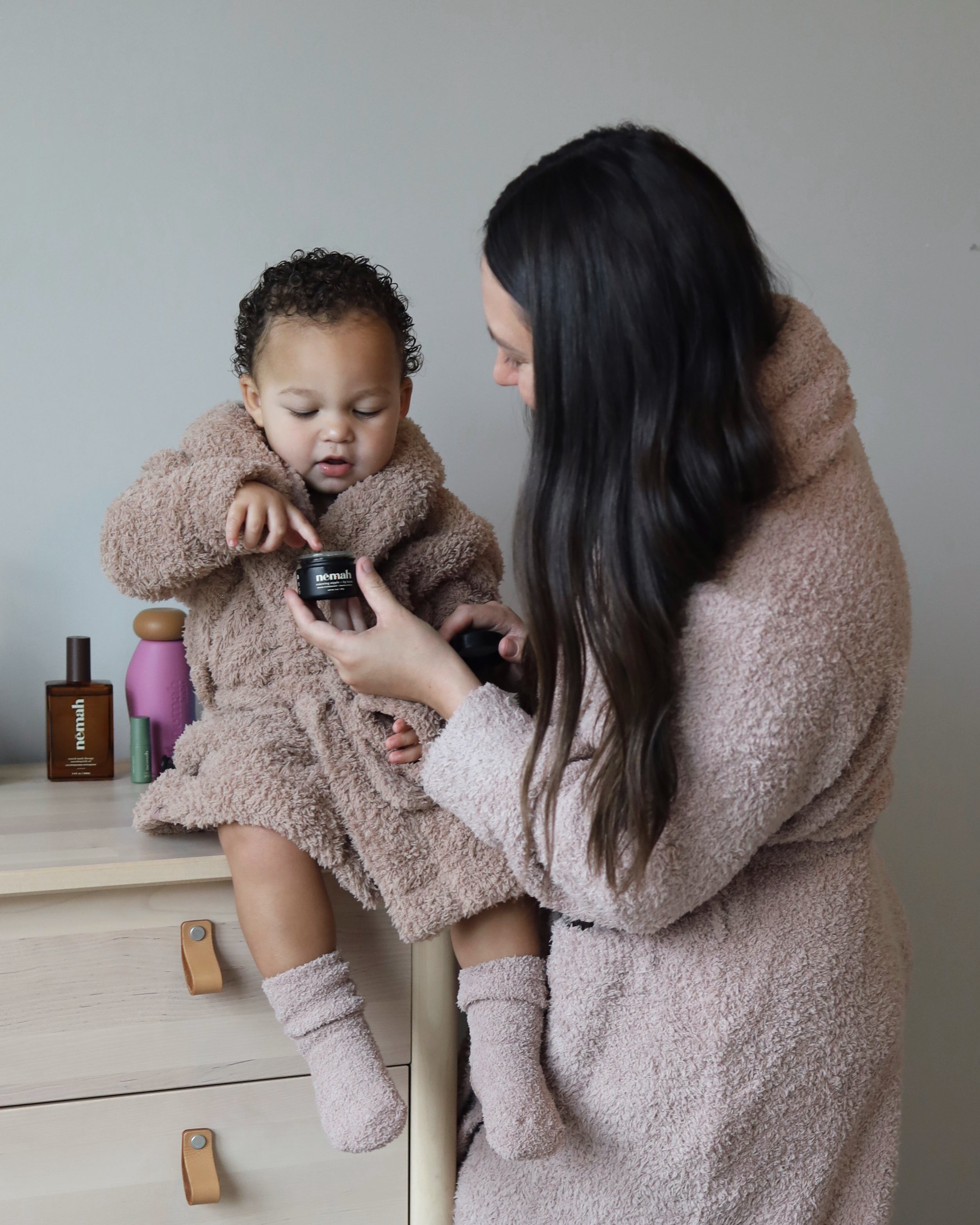 Woman showing a toddler a Nēmah jar while both wear matching fuzzy robes in a neutral indoor setting
