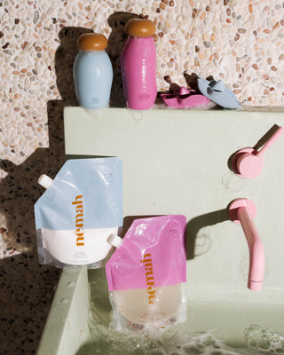 Two refill pouches labeled 'nemah' in blue and pink, two bottles with orange and pink caps, and two pairs of baby shoes in pink and blue placed on a bathtub edge with a terrazzo wall background.