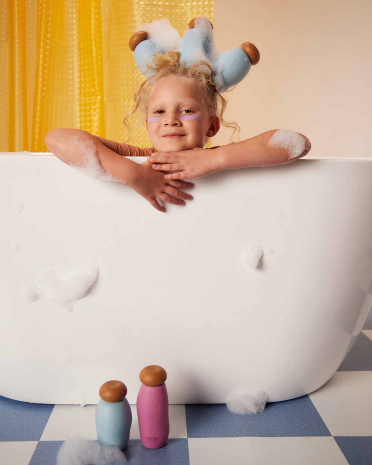 a blond child is looking from the bath tub with foam and three blue bottles on her head 