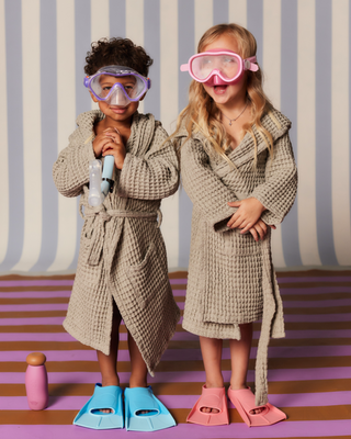 Two children wearing waffle bathrobes, snorkeling masks and colorful swim fins; one holds a snorkel, pink water bottle on the floor.