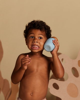 A young boy with curly hair holding a blue and brown toy phone to his ear with a surprised expression