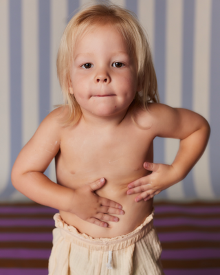 Young child with blonde hair, shirtless, standing with hands on stomach against a striped background.