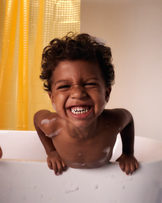 Smiling young child with curly hair leaning over the edge of a bathtub with soap bubbles on their skin and a yellow shower curtain in the background.