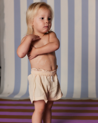 Toddler with light hair standing shirtless with hands crossed over chest against blue-and-white striped backdrop.