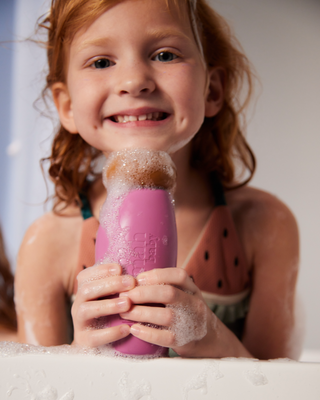 Smiling child with red hair holding a pink foamy bottle labeled "mama baby" in a bath setting