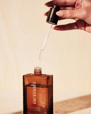 Hand holding a dropper releasing liquid above a brown bottle labeled 'nemah' with water droplets on it