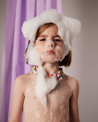 Child with soap bubbles covering head and chest, wearing a colorful beaded necklace, standing in front of a purple curtain.
