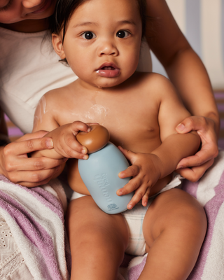Baby holding a blue bottle with a brown cap, assisted by an adult's hands, sitting on a purple and white towel.