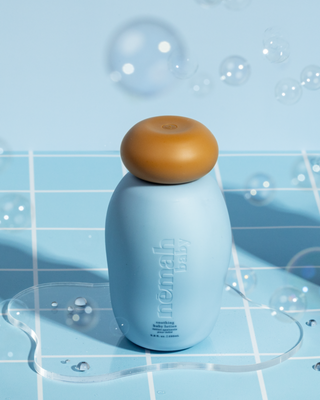 Blue Nemah baby soothing baby lotion bottle with a brown cap on a blue tiled surface with water droplets and bubbles around it