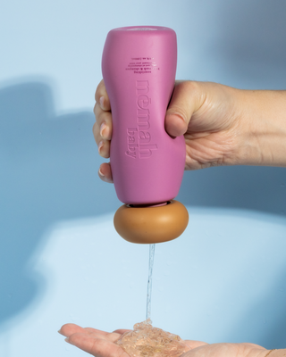 Hand squeezes upside-down pink nemah baby bottle, clear wash pouring into cupped palm.