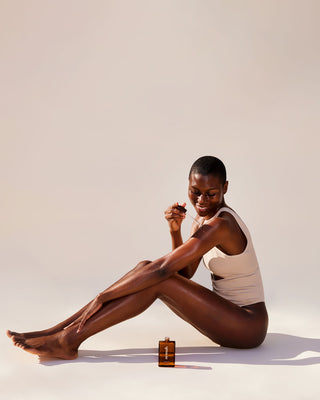Smiling woman in beige tank and underwear seated applying nemah oil from an amber bottle to her legs