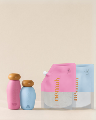Two baby product bottles, one pink and one blue, each with a brown cap, alongside two refill pouches, one pink and one blue, labeled 'némãh' with product details and '2x refill bottles' icons.