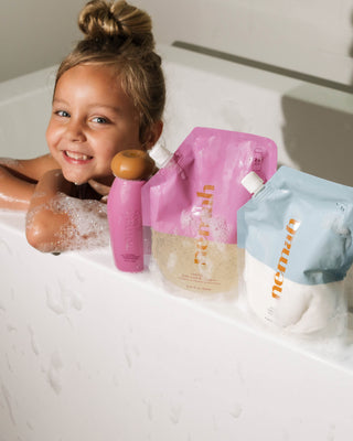 Smiling child in bathtub with pink and blue refill pouches labeled baby wash & shampoo.