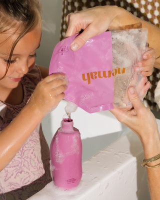 Child and adult refilling a pink bottle with a pink pouch labeled 'nomanh' containing bath wash and shampoo, with soap suds visible on hands and bottle.