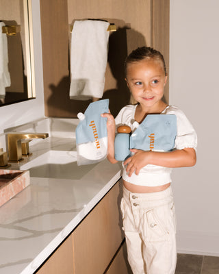 Young girl holding two blue lotion refill pouches labeled 'nemah' and a blue bottle in a bathroom setting.