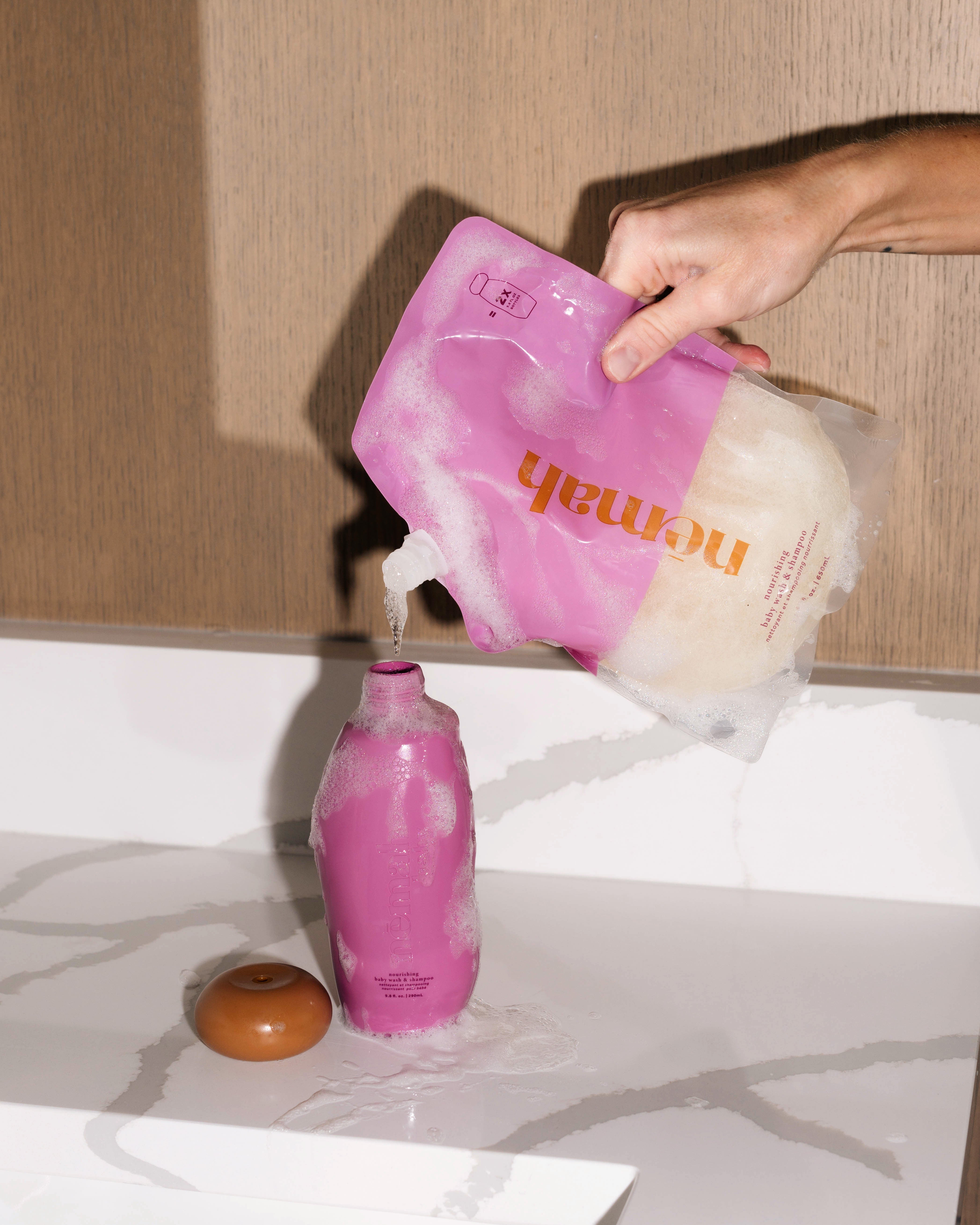 Hand pouring liquid from a refill pouch into a bottle on a counter.