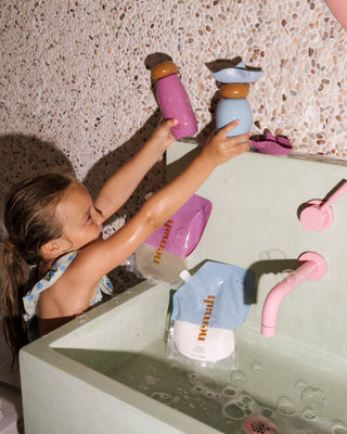 Young child reaching across tub holding pink and blue bath bottles beside refill pouches labeled 'nemah'.