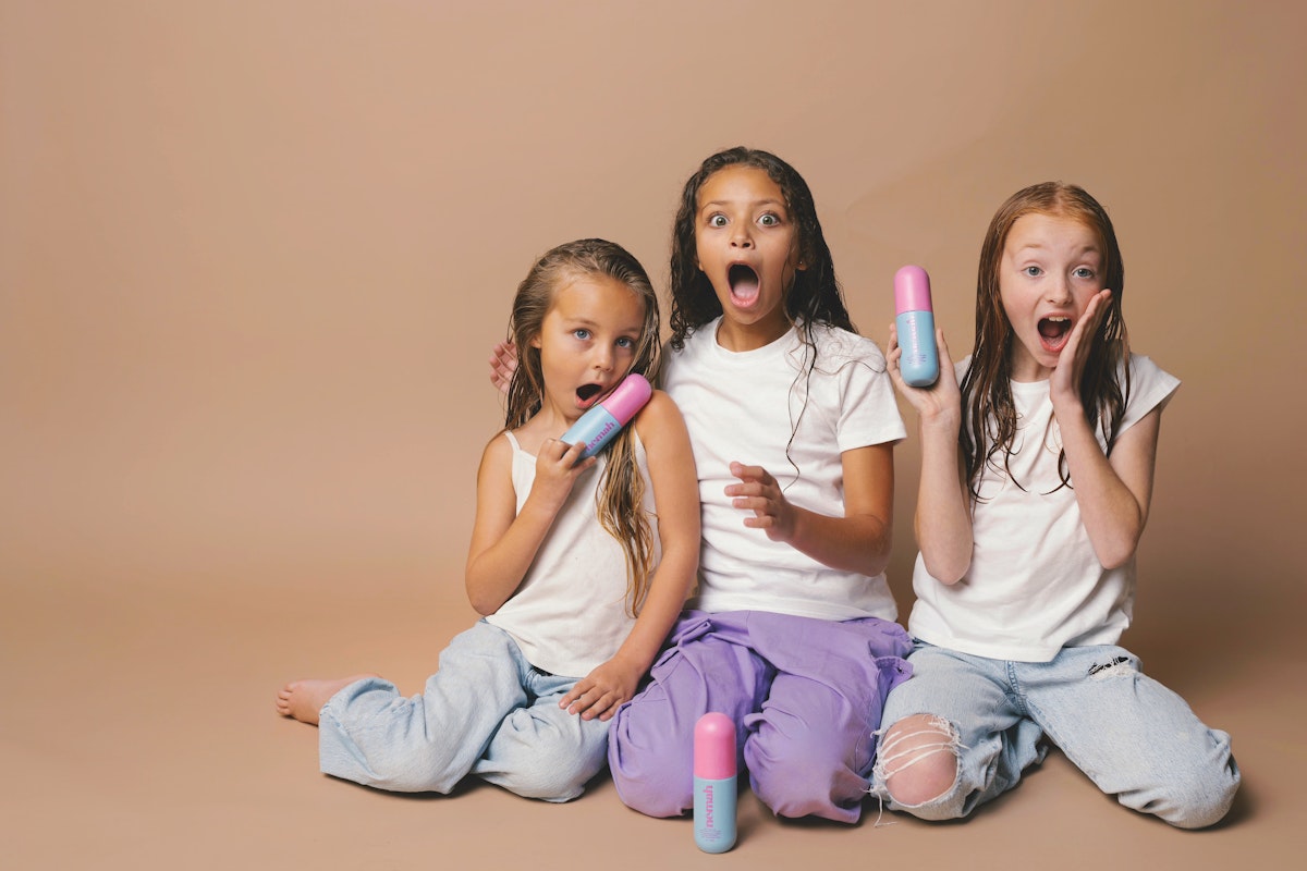 Three young girls sitting on the floor in a studio, holding colorful deodorant sticks and showing surprised expressions. They are wearing casual outfits with white tops and light-colored jeans.