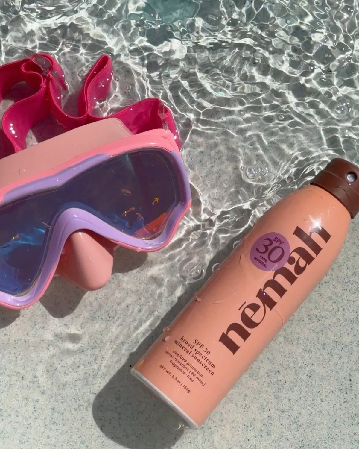 Angled view of Nēmah Kids Mineral Sunscreen Spray can showing SPF 30 sticker and water droplets beside pink swim goggles in shallow pool water