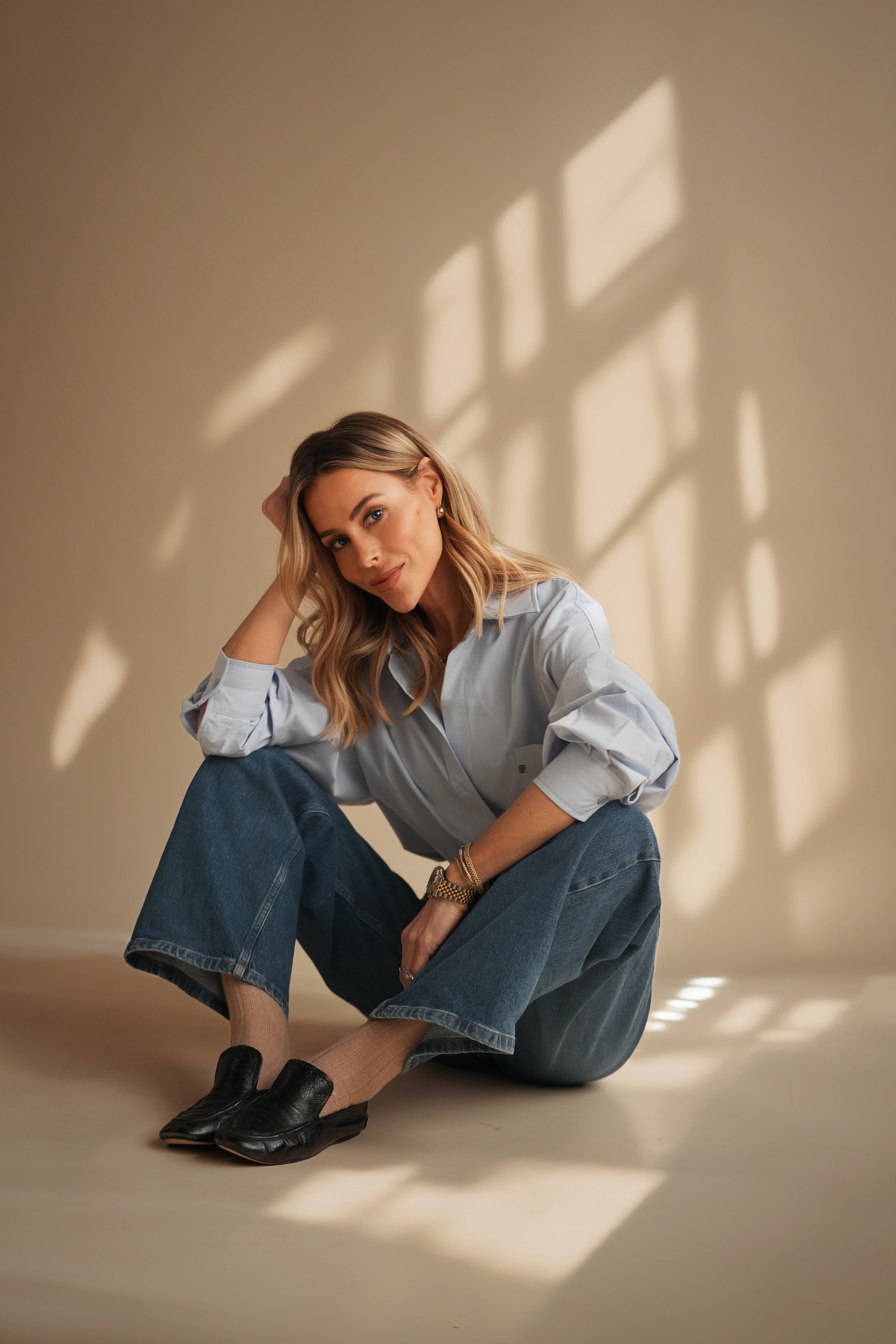 Nēmah founder seated on a studio floor wearing a light blue button-up, wide-leg jeans, and black slip-on shoes with window-pattern shadows on a beige wall