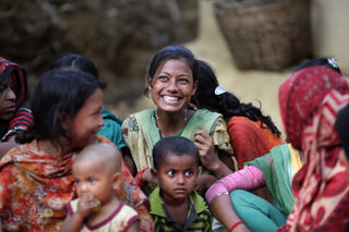 Group of people including a smiling young woman in a green dress, children, and others wearing colorful clothing, gathered closely together.